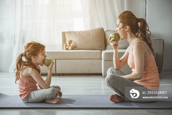 Profile of woman and kid after aerobics. They are sitting in front of each other on the floor and biting green apples with smile