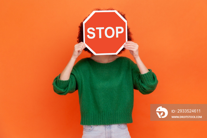 Unknown woman with wearing green casual style sweater hiding her face behind red stop traffic symbol, warning, avoids forbidden actions. Indoor studio shot isolated on orange background.