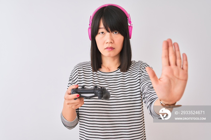 Chinese gamer woman playing video game using headphones over isolated white background with open hand doing stop sign with serious and confident expression, defense gesture