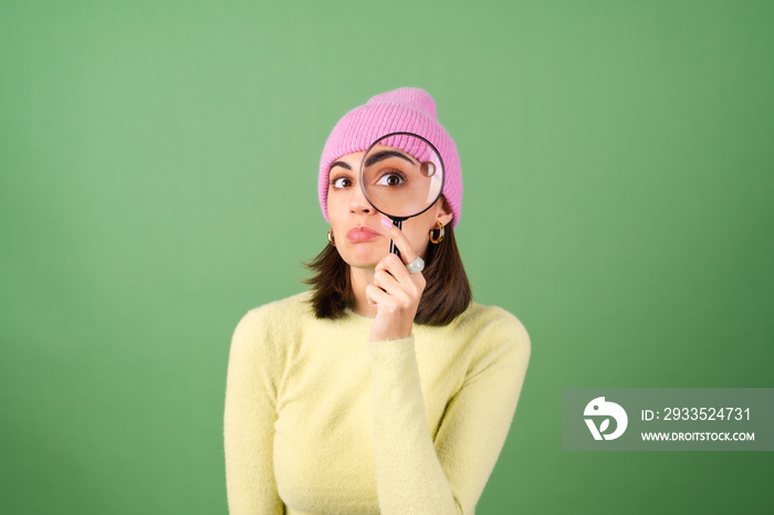 Young woman on green background in yellow sweater with magnifying glass