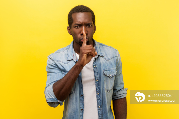 Shh, keep silence. Portrait of serious handsome man in denim casual shirt with rolled up sleeves gesturing to be quiet, asking for secrecy conspiracy. indoor studio shot isolated on yellow background