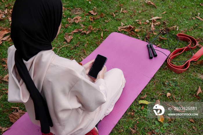 Rear view of woman in hijab holding smart phone on pink yoga mat on grass