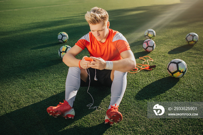 Young player sit alone on green lawn. He has rest. Guy hold phone in hands with headphones. Six balls are behind him.