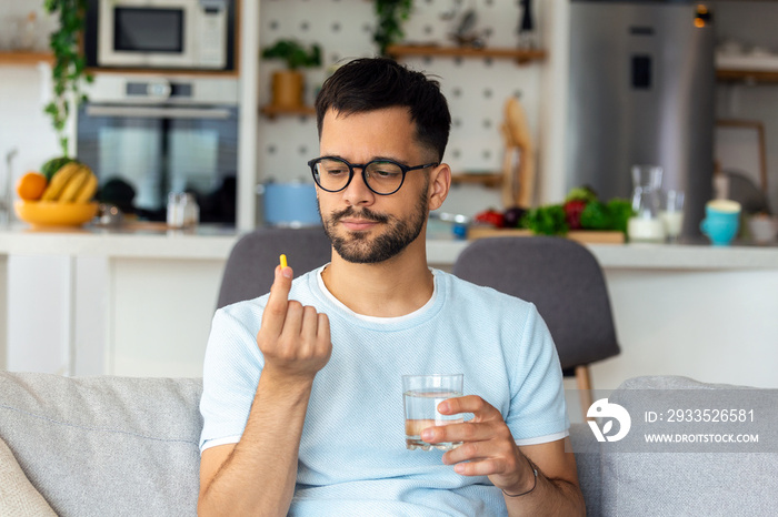 Just one pill can help. Handsome young man holding a glass of water and looking at a pill in his hand while sitting on the couch at home