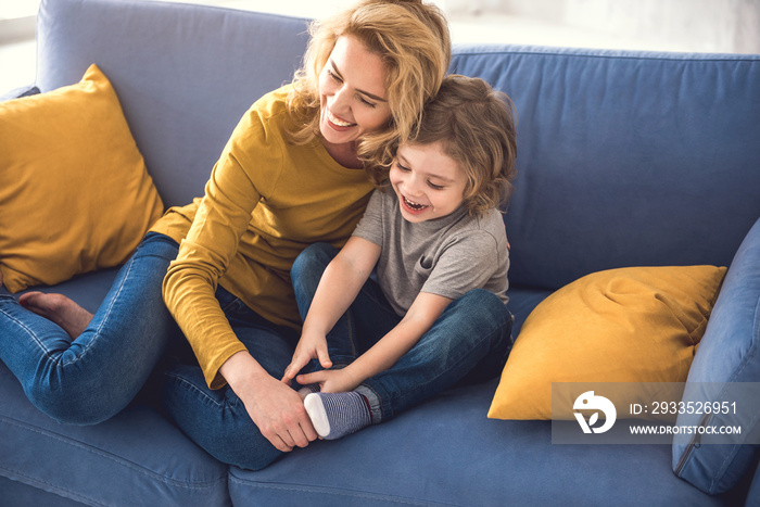 Mom and kid are laughing together in cozy home. They are holding each other with joy while relaxing on sofa. Parent is playfully touching boy feet and he is enjoying it