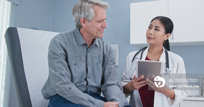 Asian woman doctor talking senior Caucasian male patient sitting on exam table. Medial professional using tablet computer to take medical history from older man