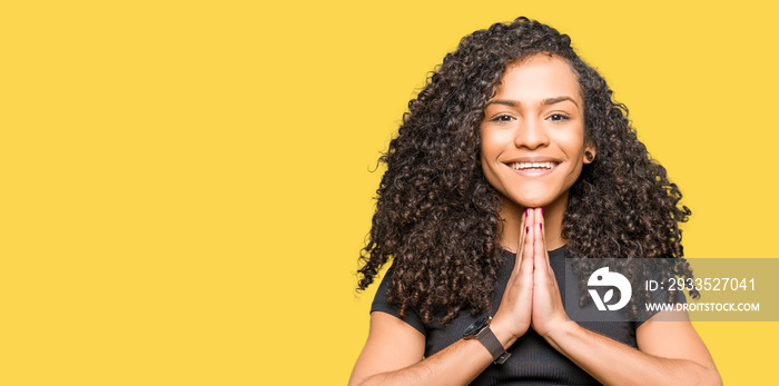 Young beautiful woman with curly hair praying with hands together asking for forgiveness smiling confident.