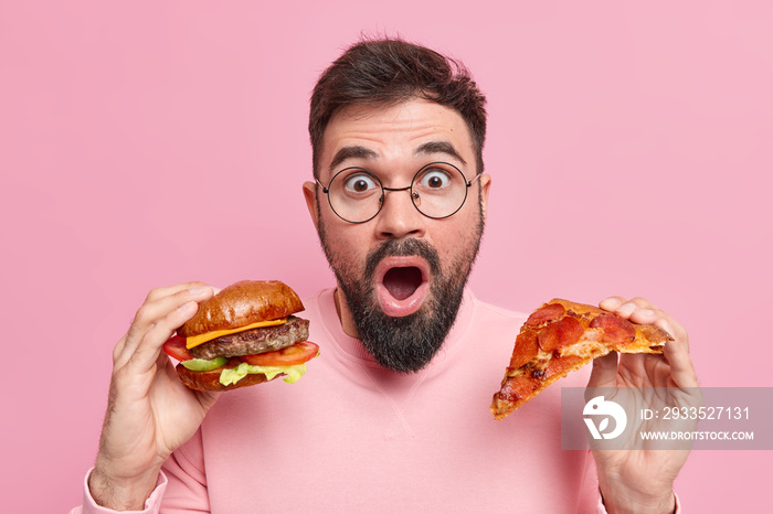 Surprised bearded man holds piece of pizza and hamburger eats junk food shocked to consume much calories wears round spectacles casual jumper isolated over pink background. Cheat meal concept