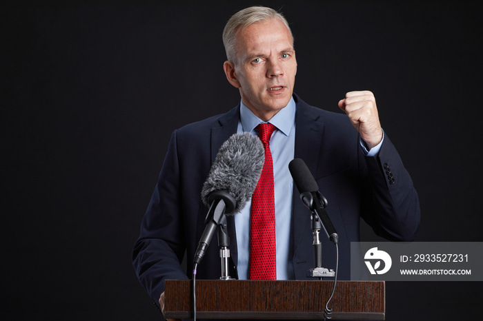 Waist up portrait of mature man standing at podium and gesturing while giving speech to microphone against black background, copy space