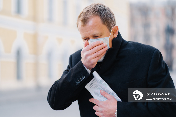 Sick man coughs, covers mouth with palm, wears medical mask, has symptoms of allergy, flu, influenza or coronavirus, walks outdoor, holds newspaper, feels unwell. Covid-19, quarantine concept