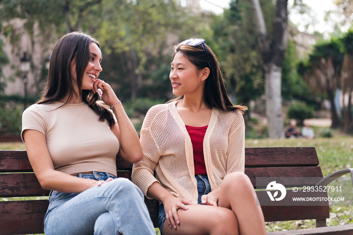 two young women chatting happy seated on a park bench, concept of female friendship and teenager lifestyle