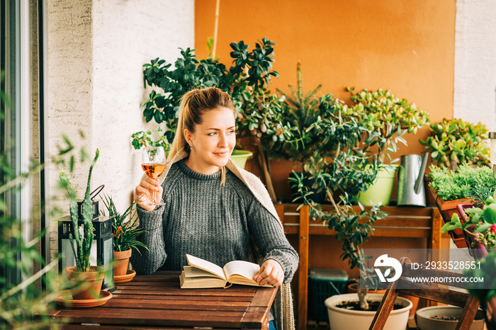 Young beautiful woman relaxing on cozy balcony, reading a book, wearing warm knitted pullover, holding glass of wine