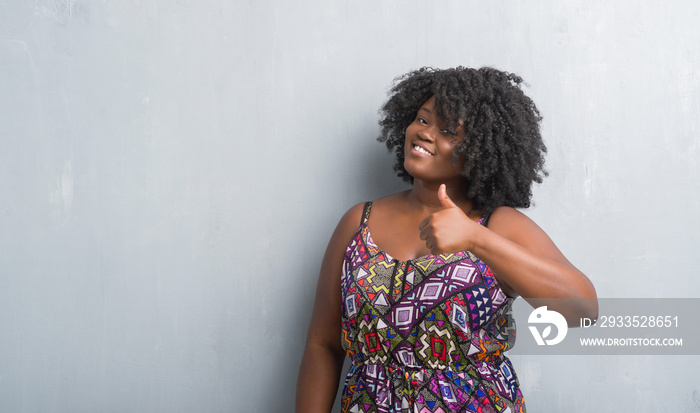 Young african american woman over grey grunge wall wearing colorful dress doing happy thumbs up gesture with hand. Approving expression looking at the camera with showing success.