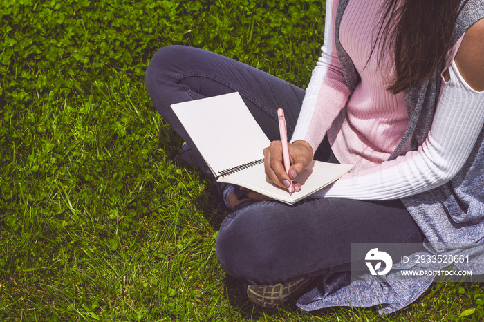 Young girl writes with pen in notebook sitting on green grass in park on meadow