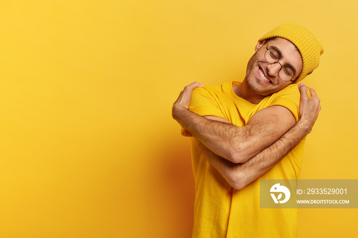 Pleased Caucasian man hugs himself, has high self esteem, tilts head, has toothy smile, wears casual yellow hat and t shirt, stands indoor, copy space for your promotional content, feels comfort