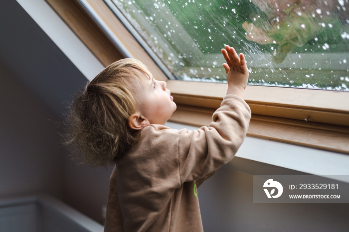 Little preschool boy staying home in bad weather and looking  with interest through window on raindrops and hail, idoors.