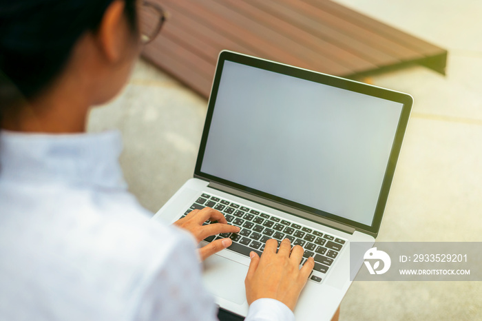 Businesswoman wearing glasses from behind view typing laptop keyboard on her lap with blank space display in front office background and sun flare effect- woman office worker- businesswoman concept