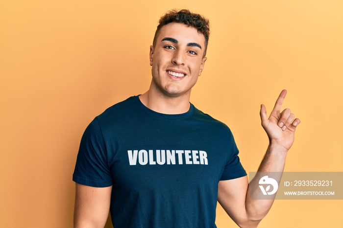 Hispanic young man wearing volunteer t shirt cheerful with a smile on face pointing with hand and finger up to the side with happy and natural expression