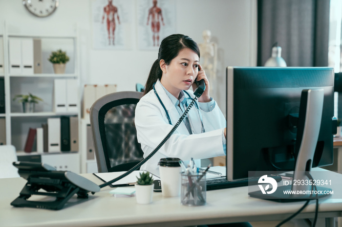 Serious asian female doctor on phone in office. young japanese woman medical worker in clinic hospital connecting with nurse on telephone call checking patient situation of surgery on computer screen