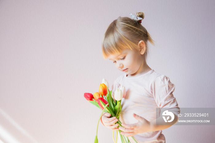 A serious little blond-haired girl is holding a bouquet of colorful tulips in hands in a sunny room at home. Holy concept.