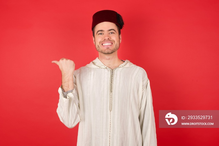 Close up photo of lovely young caucasian muslim man wearing djellaba and traditional hat over red background pointing aside with forefinger, showing at copy space having news about bargains