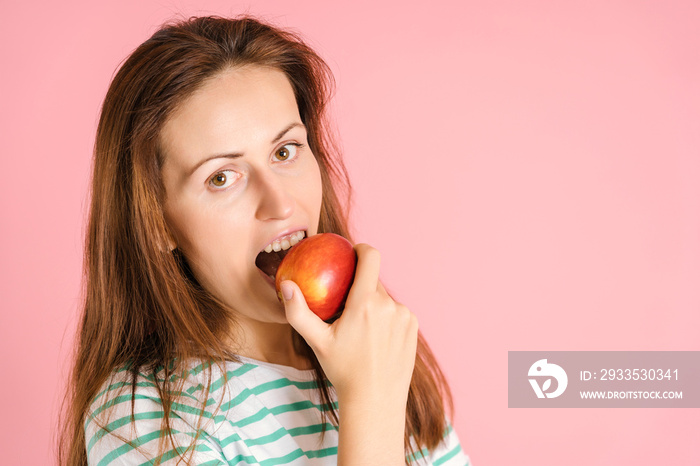 Portrait of a middle-aged woman biting a red apple on a pink background