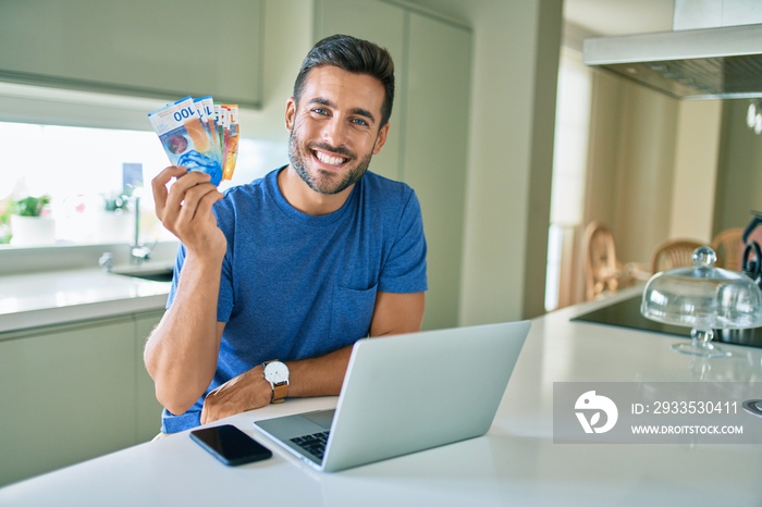 Young handsome man smiling happy holding swiss franc banknotes at home