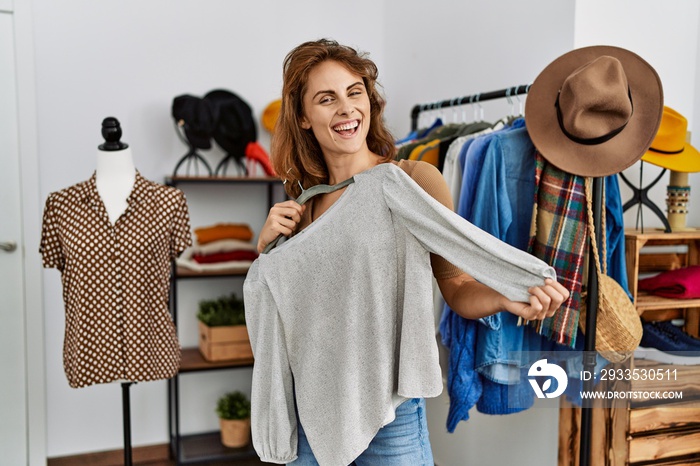 Young caucasian customer woman smiling happy holding hanger with clothes at clothing store.