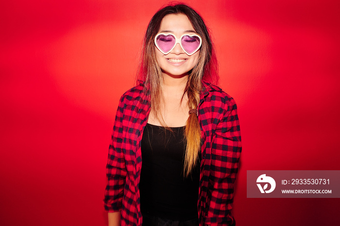 Portrait of a cheerful Asian Kazakh student girl in a plaid shirt and funny glasses laughs and smiles isolated on a red studio background