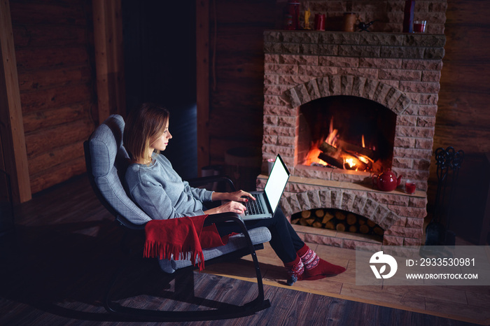 Cozy home. Pretty young woman working on laptop computer near the fireplace.