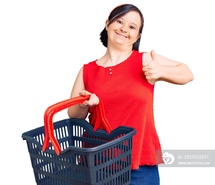 Brunette woman with down syndrome holding supermarket shopping basket smiling happy and positive, thumb up doing excellent and approval sign