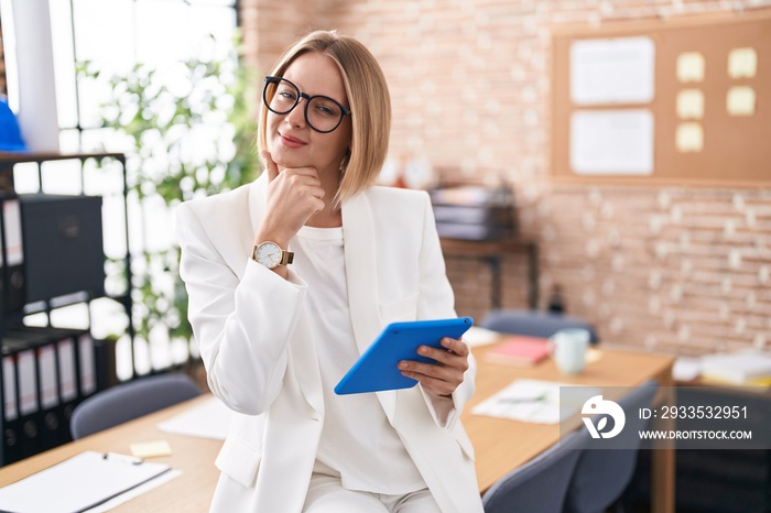 Young caucasian woman working at the office wearing glasses looking confident at the camera smiling with crossed arms and hand raised on chin. thinking positive.