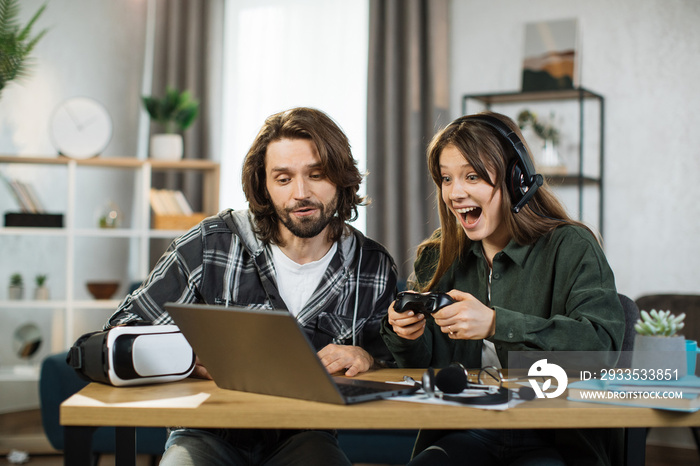 Portrait of excited focused young girl in headset playing in video game using laptop and joystick. Bearded man, sitting near woman, cheering her for victory. Happy leisure time, game addiction