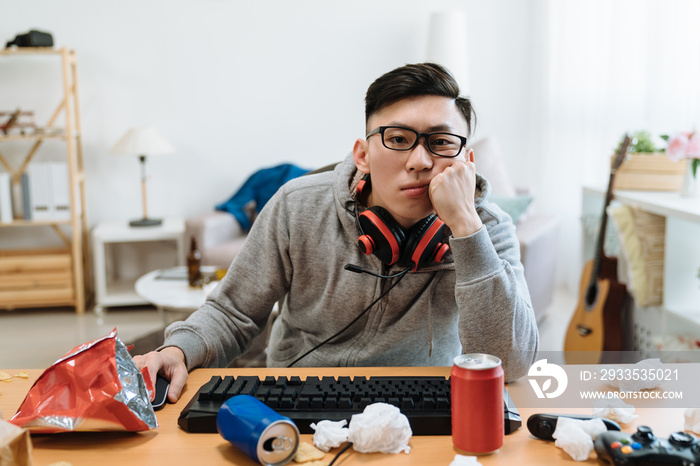 Bored guy gamer playing video online games on computer while sitting by messy table at home. lazy asian man with headsets unhappy in dirty bedroom. young male indoorsy stay indoors on hot summer day