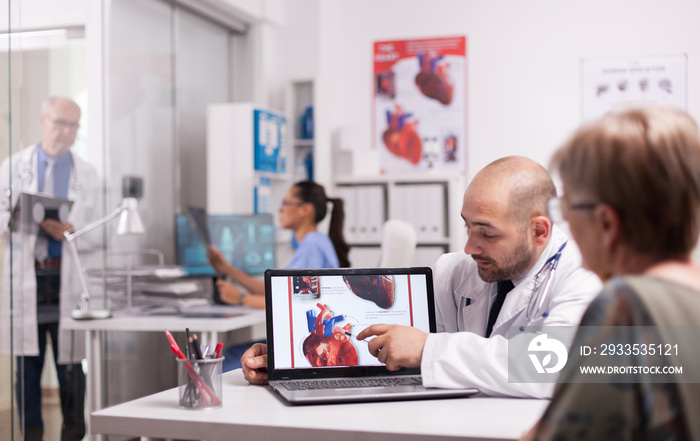 Young doctor pointing at heart on laptop screen in hospital office. Cardiologist with senior patient during consultation. Mature medic taking notes on clipboard on clinic corridor.