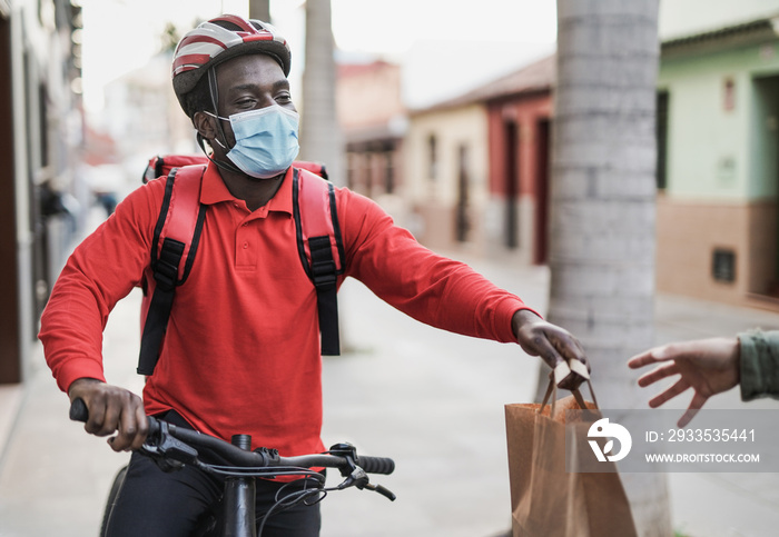 African man working for food delivery with bicycle and paper bag wearing surgical face mask for coronavirus outbreak - Food delivery occupation with safety measures