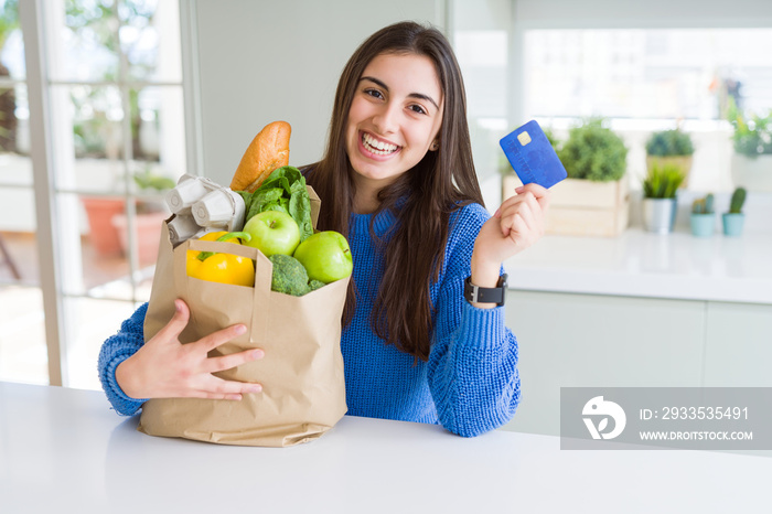 Young woman holding a paper bag full of fresh groceries and showing credit card as payment metod