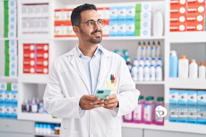 Young hispanic man pharmacist using smartphone working at pharmacy