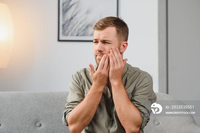 Dental health problems concept. Man suffering from strong toothache. Upset male patient with inflamed nerve, tooth abscess, or teeth sensitivity sitting on sofa and touching cheek with grimace of pain