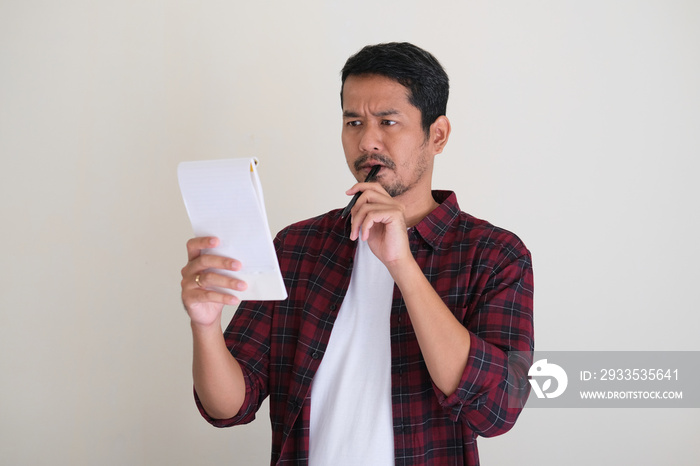 Asian man bite a pen while reading notes showing confused expression