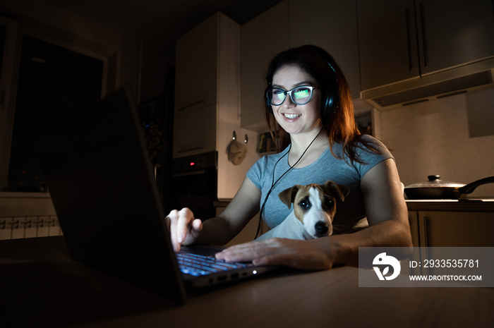 A smiling woman in headphones sits in the dark at a wireless computer in the kitchen with a puppy of Jack Russell Terrier on her knees. Girl with her little dog watching a movie on a laptop at home.