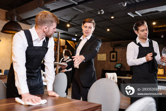 Serious displeased handsome young restaurant manager in formal jacket gesturing hand while giving task to waiter during cleanup