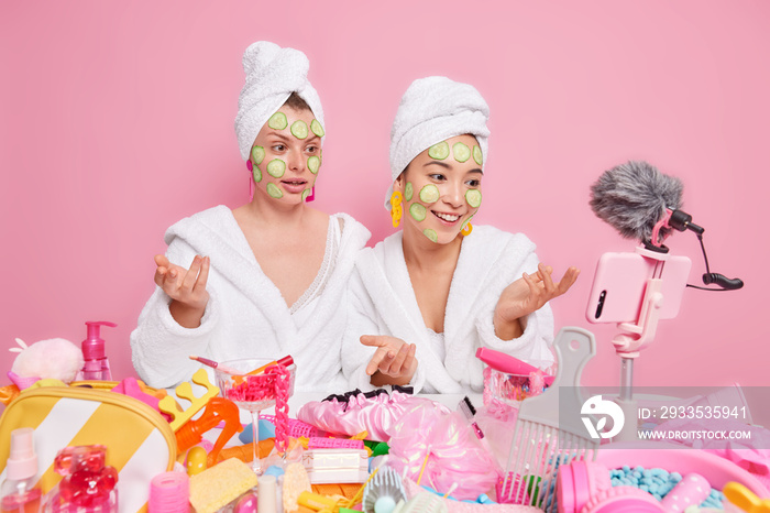 Two diverse women record tutorial video tell followers how to do beauty treatments and care about skin apply cucumber slices on face stand next to each other in bathrobes near table with cosmetics