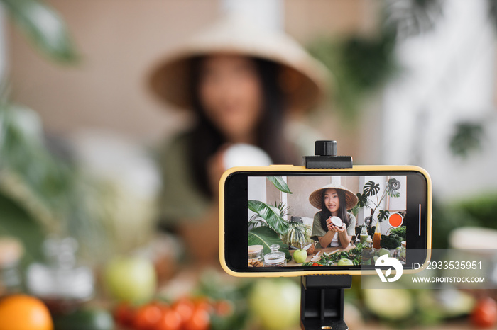 Focus on smartphone. Beauty asian blogger in traditional conical hat using coconut to make smoothies for breakfast and recording video for online channel live.