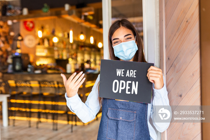 Happy female waitress with protective face mask holding open sign while standing at cafe or restaurant doorway, open again after lock down due to outbreak of coronavirus covid-19