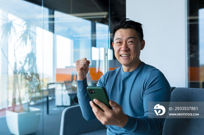 Successful and happy asian businessman man celebrating victory sitting and working in modern office at desk, celebrating victory looking at camera and joyfully shouting holding mobile phone