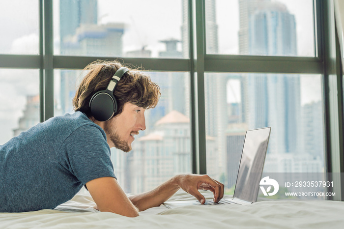 Young man is working on a laptop in his bed on a background of a panoramic window overlooking the skyscrapers. Uses wireless headphones. Freelancer, remote work, work from home