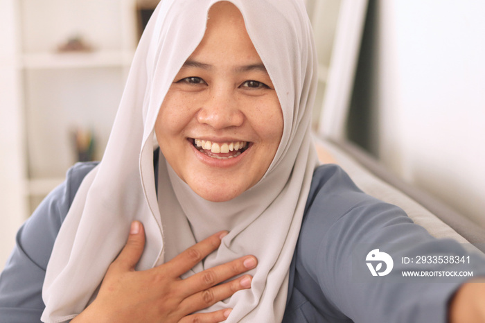 Happy cheerful beautiful Asian muslim lady wearing hijab waving to camera, doing selfie or video call gesture