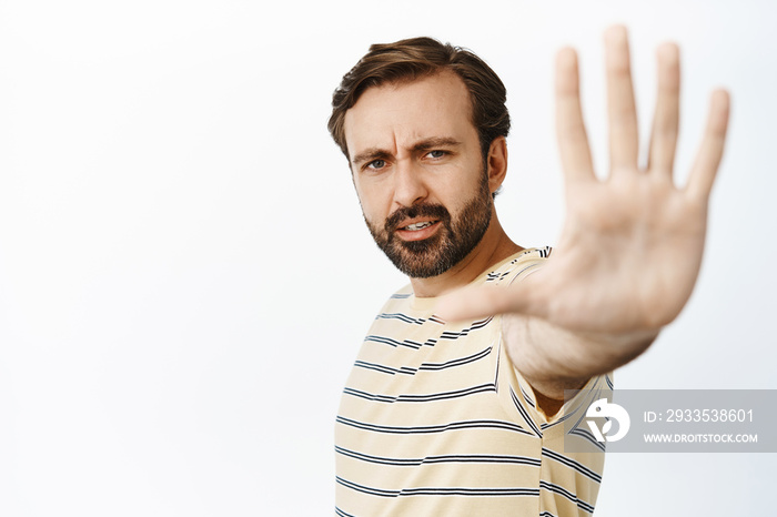 Portrait of serious guy extends his hand to stop you, showing palm taboo gesture, prohibit smth bad, standing over white background, expresses dislike