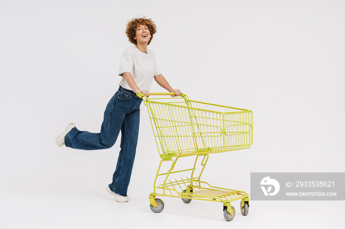 Cheerful middle-aged woman standing with shopping cart isolated over white background